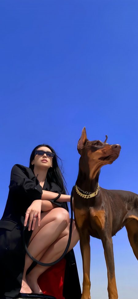 美女黑衣墨镜蹲姿与杜宾犬金链配饰蓝空下人物摄影手机壁纸